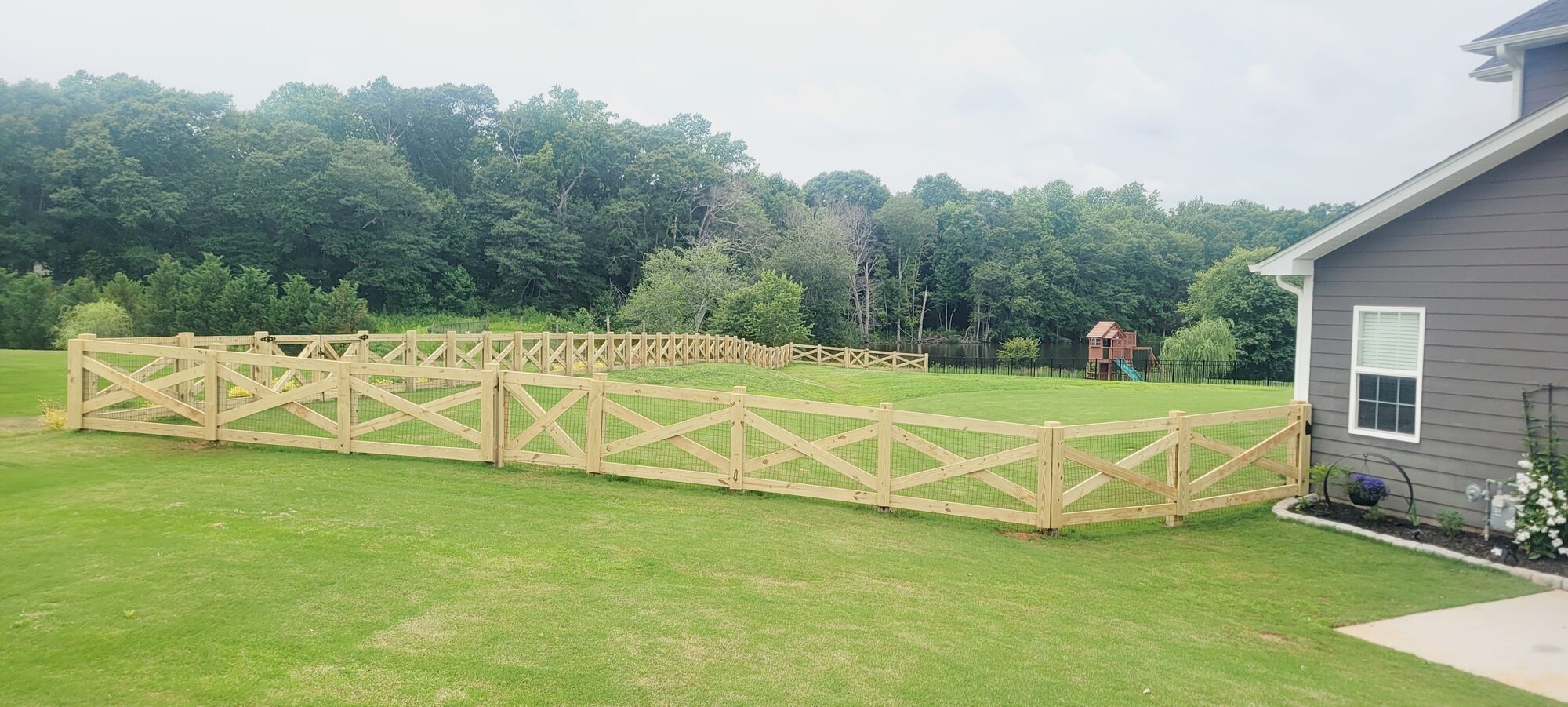 A natural, unstained wood rail fence around a backyard with a play set and pond